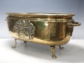 A two handled brass Jardiniere, oval and with embossed coat of arms on four paw supports, 2ft W
