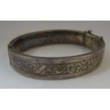 A silver bangle. Hinged oval bangle decorated with engraved scrolling foliage. With safety chain. (
