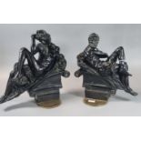 Pair of black moulded composition figures in Grecian style reclining on scrolled arches, labelled '