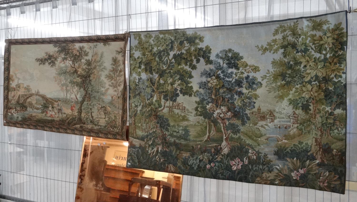 Two modern tapestry wall hangings, one of figures near a river with donkey and bridge the other a