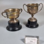 Two small silver two handled trophy cups, both on ebonised bases (one fixed). (B.P. 21% + VAT)