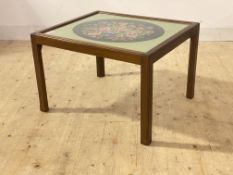 A mid century mahogany framed coffee table, the glass top with needlework panel behind, raised on