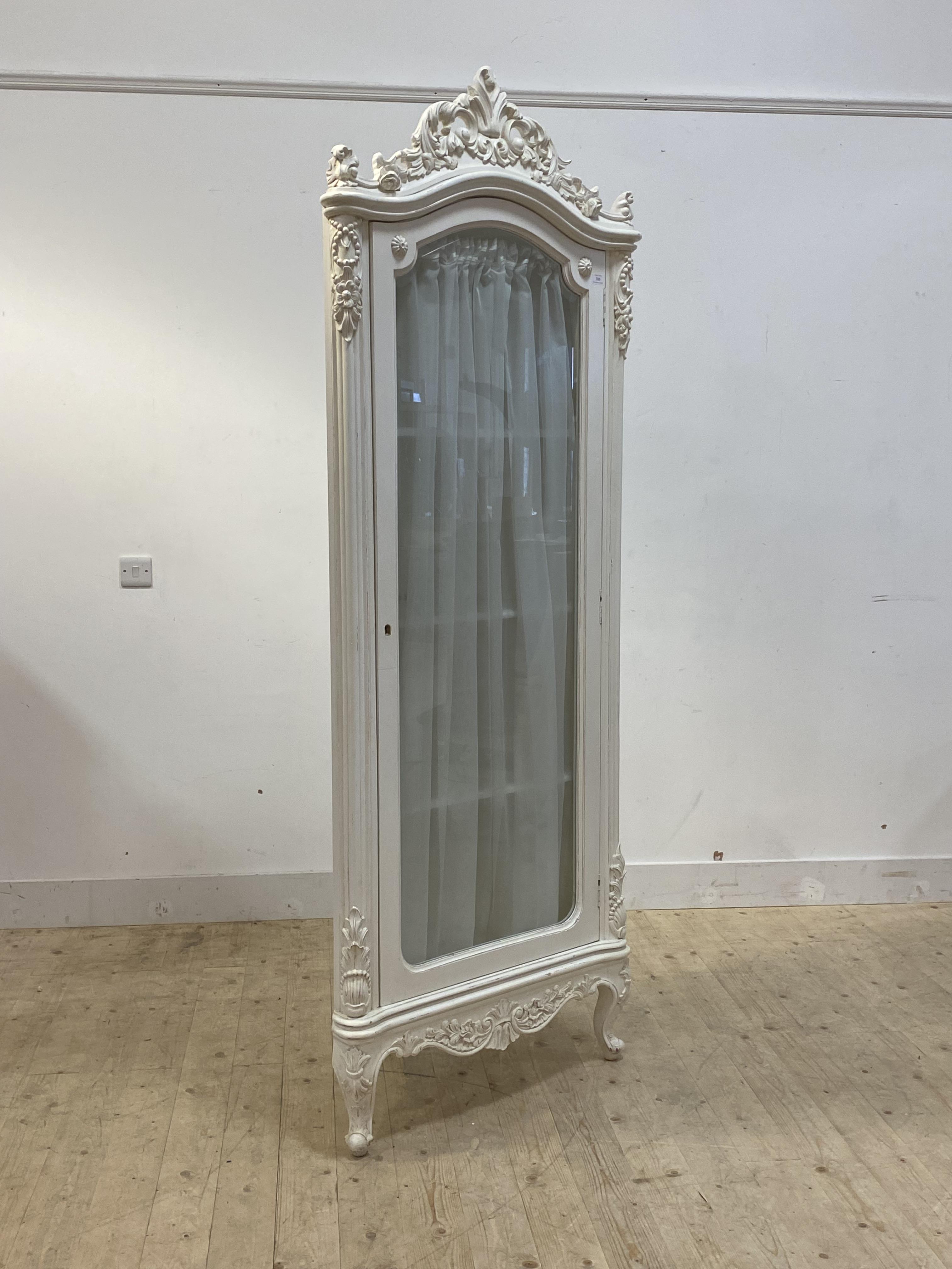 A French style white painted floor standing corner cabinet, with floral carved pediment over full