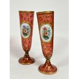 A pair of 19thc cranberry glass tapered cylinder vases with white enamelled panels depicting