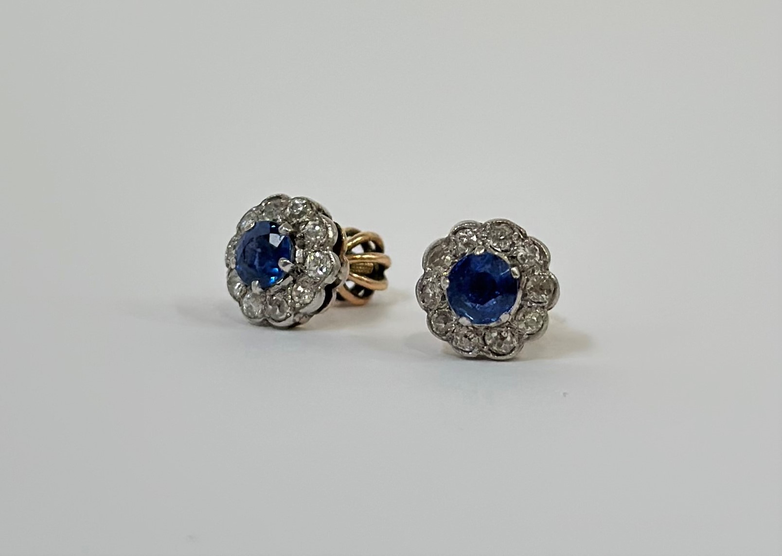 A pair of sapphire and diamond stud earrings, each with round-cut sapphire claw-set within a band of