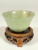 A Chinese Jadeite pale green celadon cup on raised foot and associated hardwood carved wood stand. (