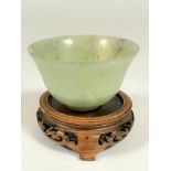 A Chinese Jadeite pale green celadon cup on raised foot and associated hardwood carved wood stand. (