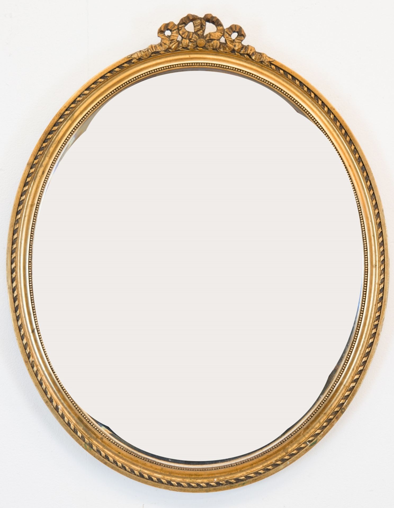 Spiegel, oval, im goldenem Holzrahmen mit Reliefdekor und Schleifenbekrönung, mit facettiertem Glas
