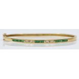 A 14K gold emerald & diamond hinged bangle, channel set with brilliant cut diamonds and square cut