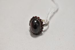 An oval garnet cabochon ring with garnet chip surround in a white metal collared mount on white