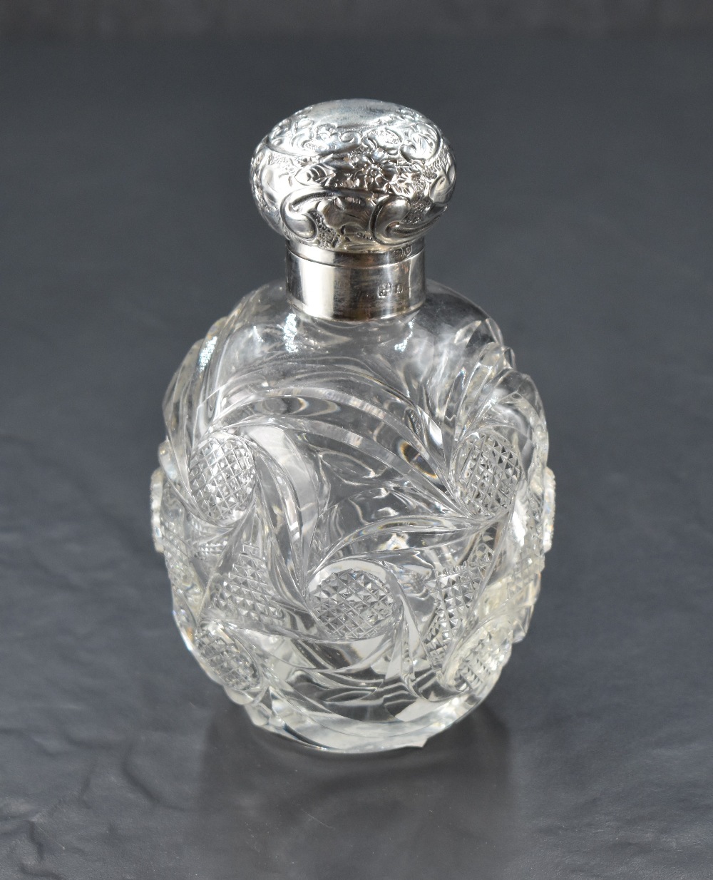 A large silver-mounted cut-glass scent bottle, having a foliate embossed and hinged cover over the