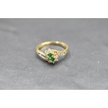 An emerald and diamond double cluster ring having diamond set cross over shoulders on an 18ct gold