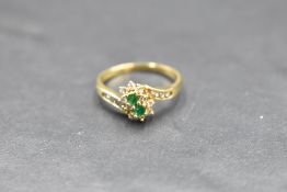 An emerald and diamond double cluster ring having diamond set cross over shoulders on an 18ct gold