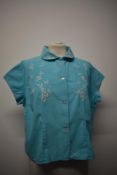 A 1950s blue blouse, having white embroidery, cap sleeves and button to front, medium size.