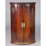 A Georgian mahogany bow-front hanging corner cabinet fitted three shelves enclosed by a pair of