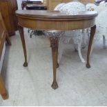 A 1930s mahogany demi-lune card table, raised on C-scrolled and foliate carved, tapered legs,