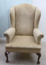 An early 20thC Georgian style wingback armchair, upholstered in mushroom coloured fabric, raised