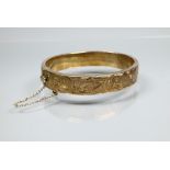 A silver-gilt half-hinged oval bangle with foliate engraved decoration, and safety chain attached,