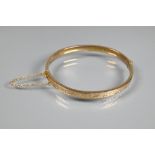 A 9ct yellow gold half hinged oval bangle, with engraved scroll decoration to one side, safety chain