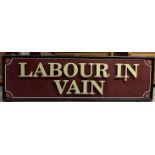 A large sign with a black frame and maroon ground, with raised letters "LABOUR IN VAIN",