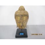 A gold painted buddha's head with green, silver and blue sequin detail on a black stand Location: