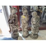 A group of three weathered concrete garden statues depicting maidens in various poses, the tallest