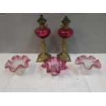 A pair of cranberry and brass oil lamps with embossed decoration, along with a pair of light shades,