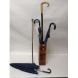A group of vintage umbrellas, a walking stick, and a swagger stick in a wooden stick stand to