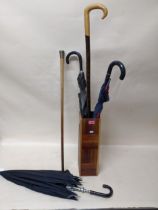 A group of vintage umbrellas, a walking stick, and a swagger stick in a wooden stick stand to