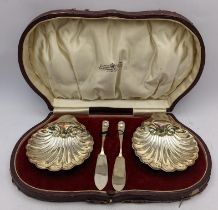 A pair of early 20th century silver butter dishes of shell design, together with two knives, in a