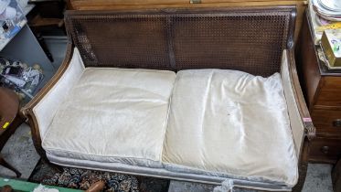A 1920s cane two-seater sofa with loose cushions, castors missing, ram's head arms and blind