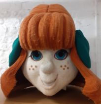 A Carnival head in the form of a cartoon female having orange woollen hair and a large green bow,