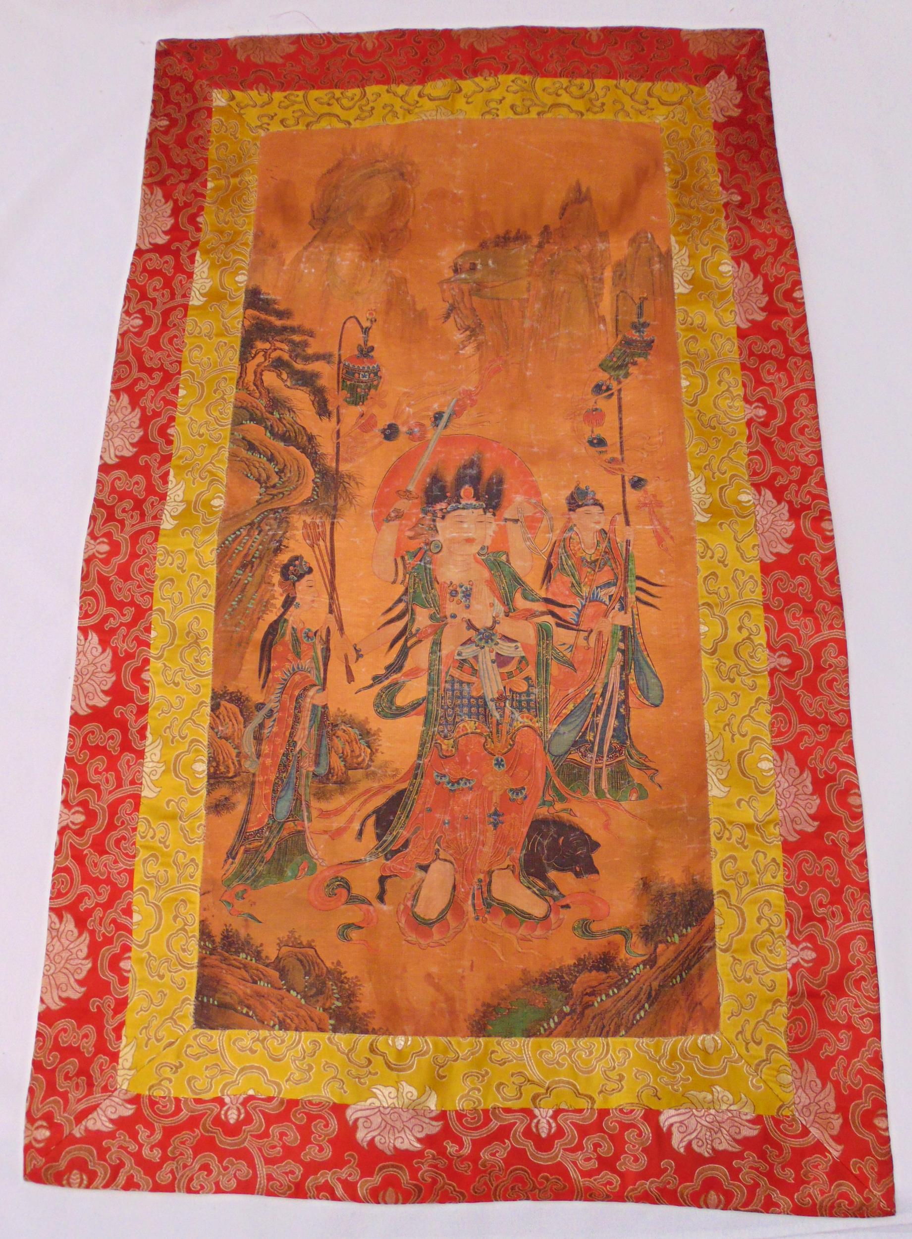 A Tibetan silk Tanka decorated with figures in a landscape within a red silk border, 67 x 111cm