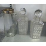 A PAIR OF CUT GLASS SQUARE SHAPED DECANTERS AND STOPPERS AND A CLEAR GLASS TALL JUG WITH SILVER