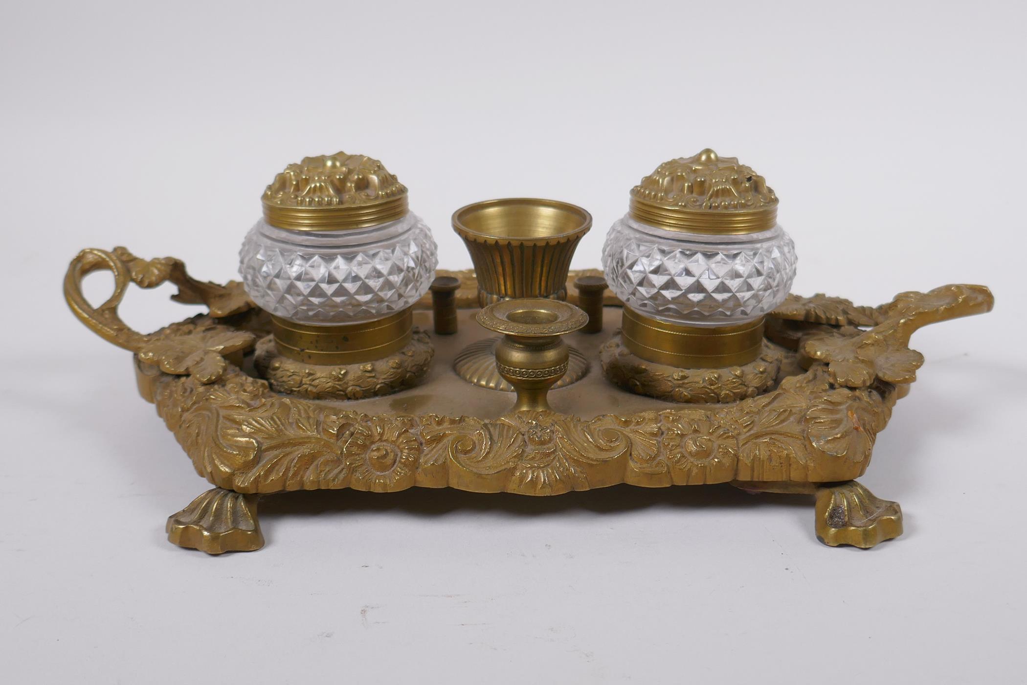 An antique brass desk stand with raised vine decoration and two glass inkwells, 30 x 20cm