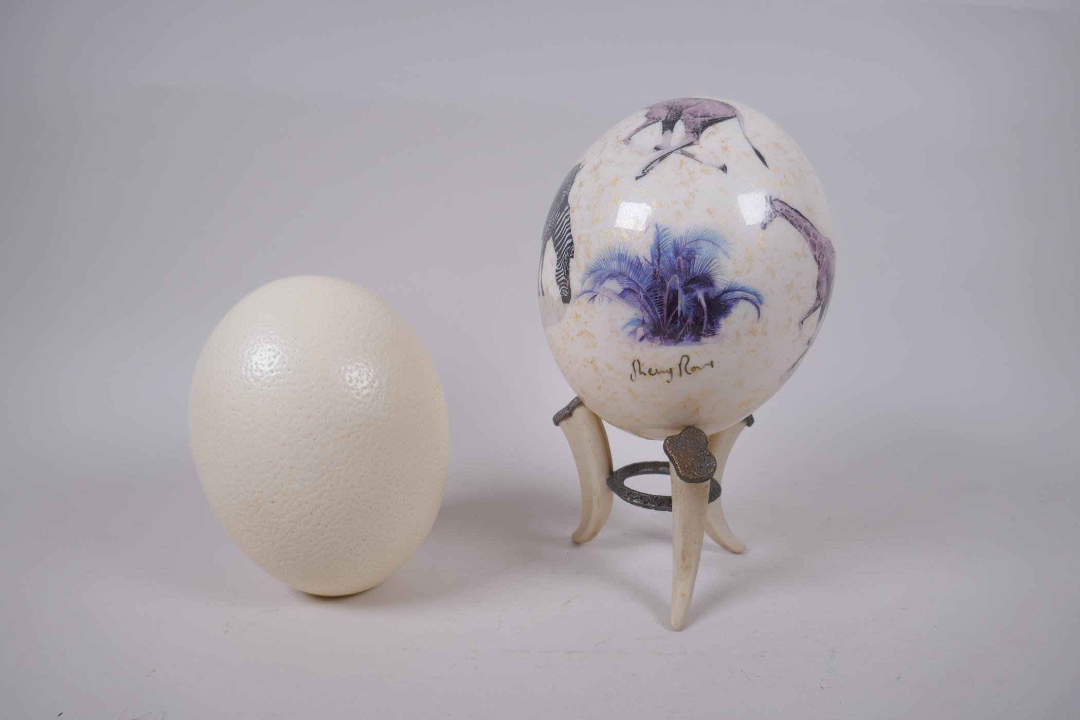 A decorative gilt and collage ostrich egg on stand by Sherry Rowe, and another undecorated ostrich