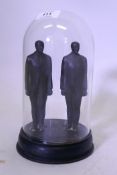 A pair of cast iron figures of standing men, mounted on a wood base and under a glass dome, 20cm