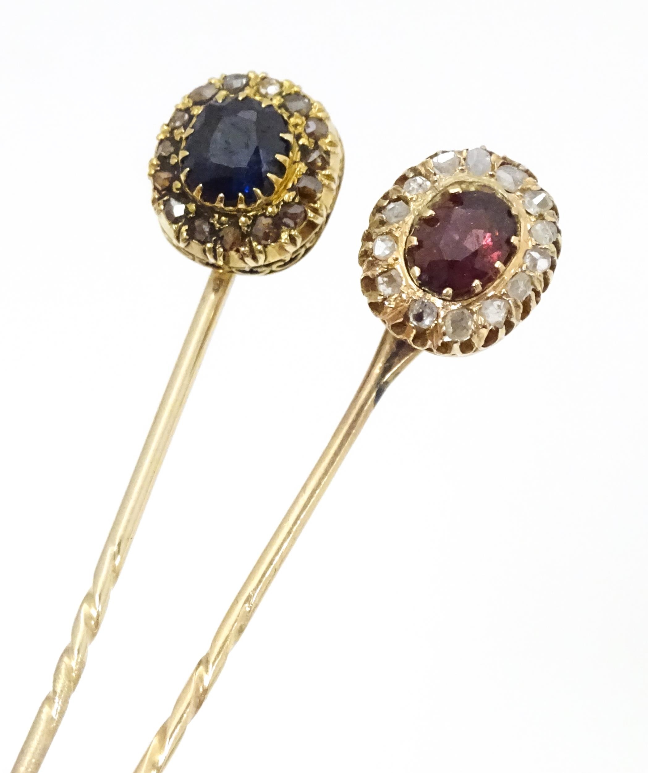 Two yellow metal stick pins, one surmounted by a central sapphire bordered by diamonds, the other