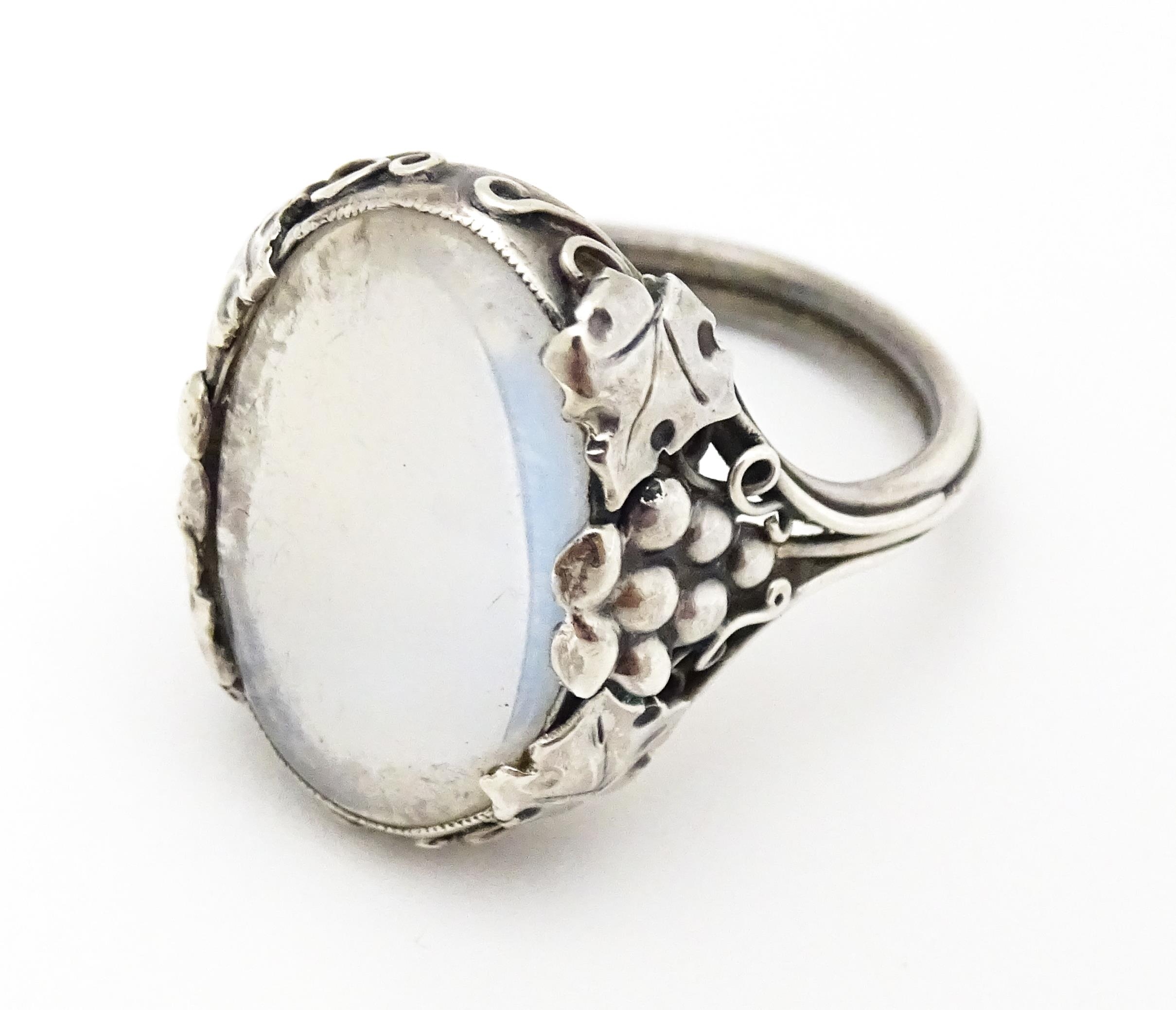 A white metal ring set with central oval moonstone cabochon and with fruiting vine detail to