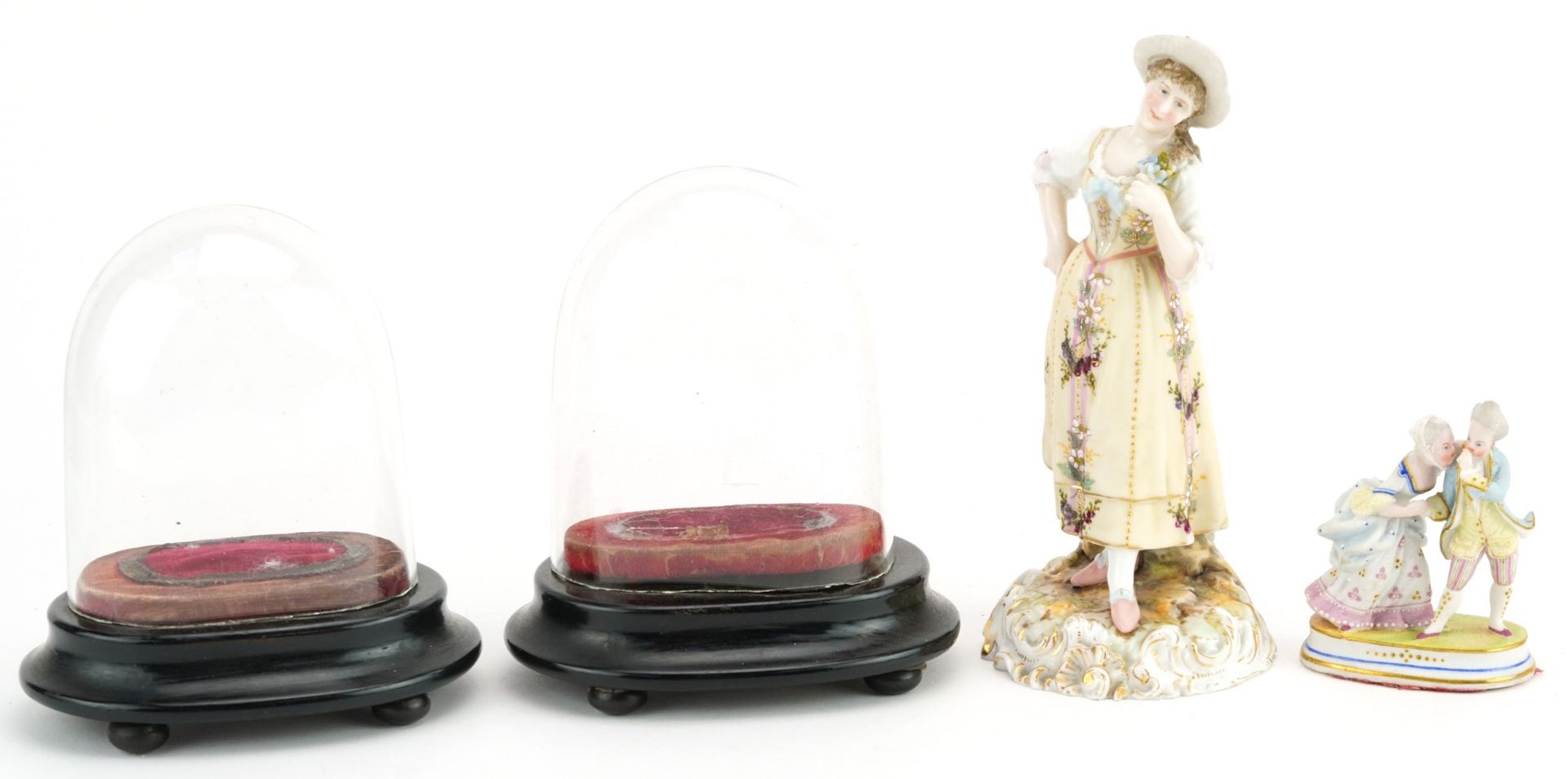 Two continental porcelain figures and a pair of glass domes raised on ebonised bases, the largest