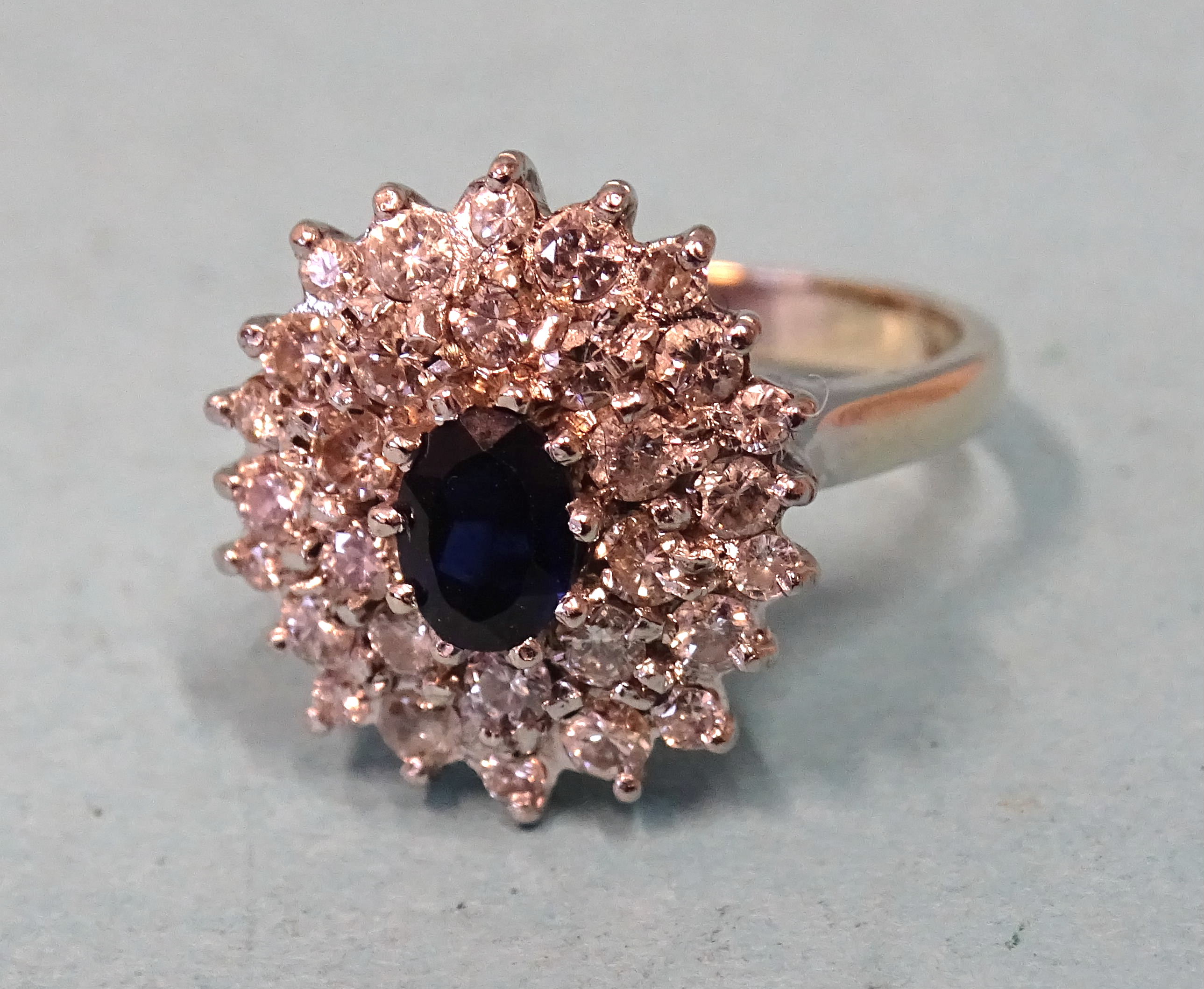 A sapphire and diamond cluster ring claw-set an oval sapphire within two tiers of a total of