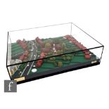 A diorama of a rural village scene with houses, roads and trees, contained in an acrylic case,