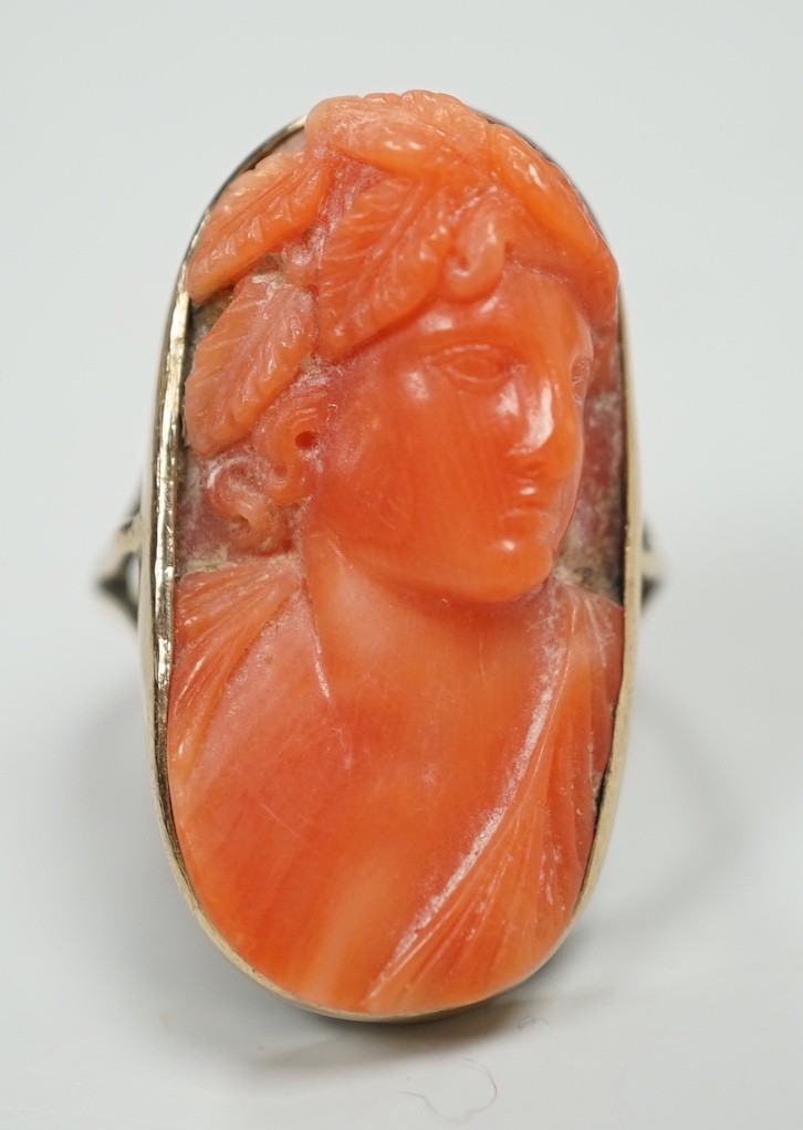 A yellow metal and coral set oval dress upfinger ring, the coral carved with a classical bust to