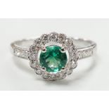 A modern 18ct white gold, emerald and diamond set circular cluster ring, with diamond set shoulders,
