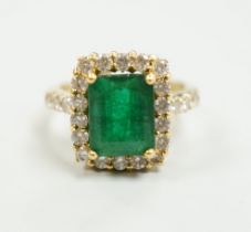 A modern 18ct gold and single stone emerald set dress ring, with diamond set border and shoulders,