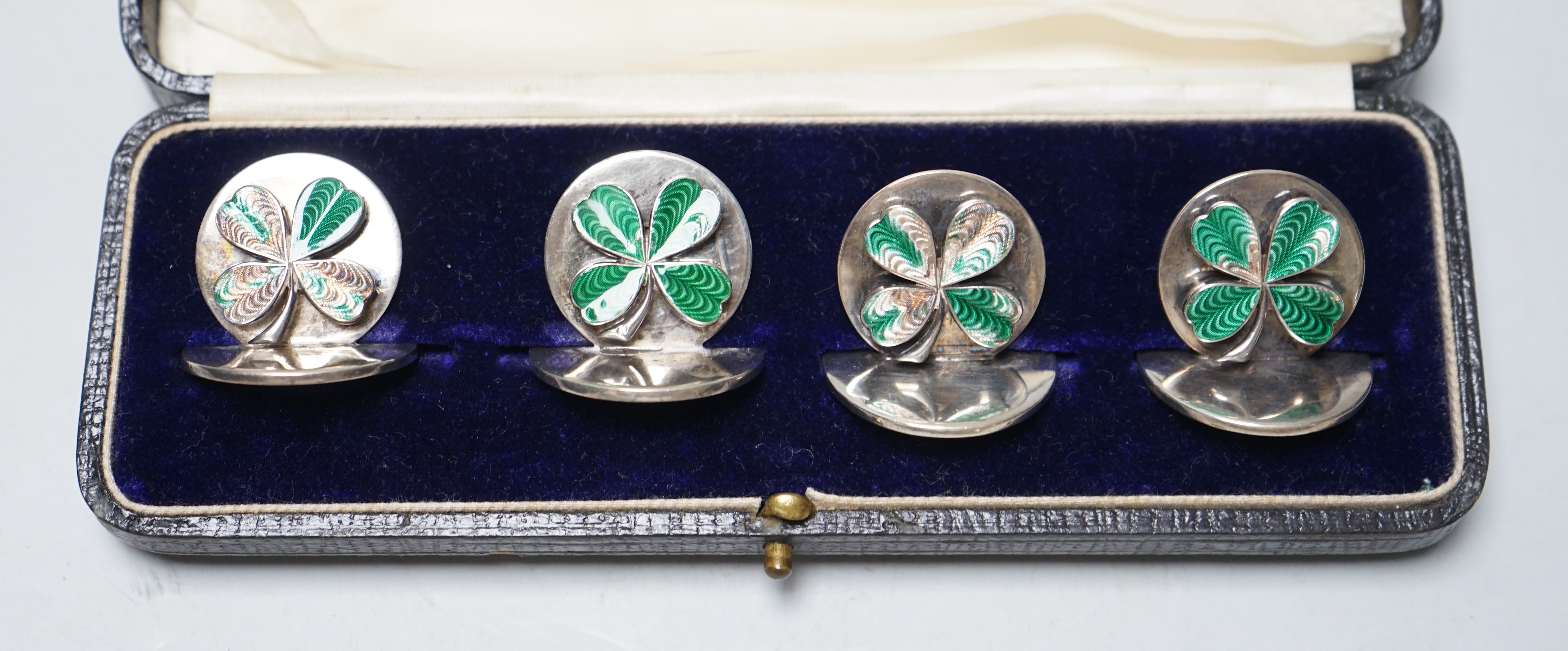 A cased set of four George V enamelled silver menu holders, decorated with four leaf clovers, by