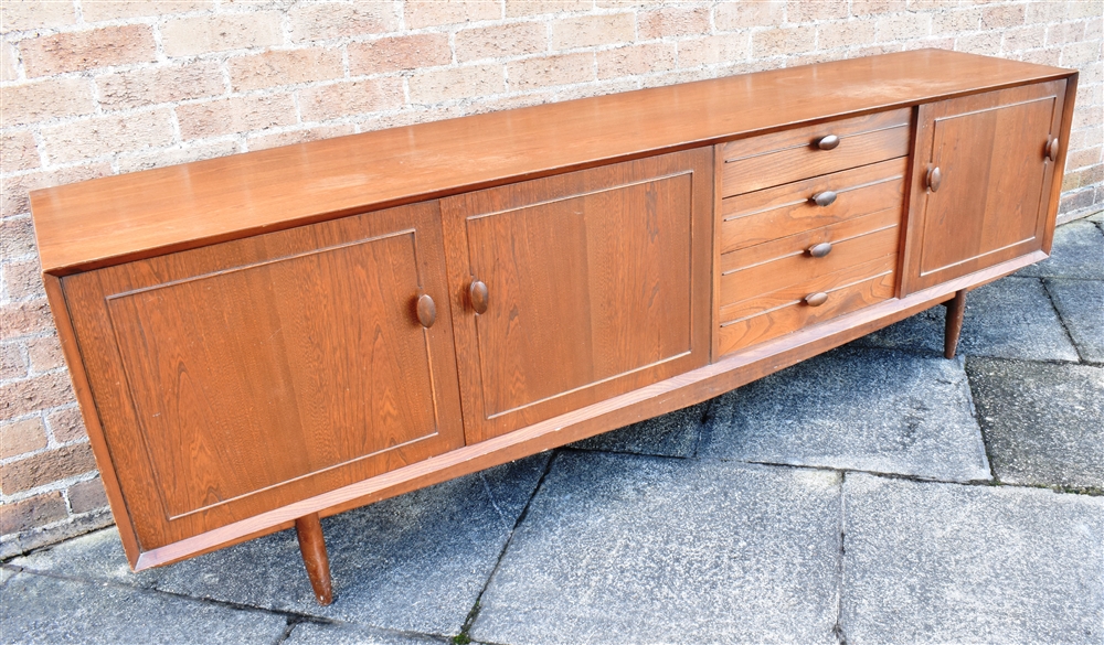 A 1960S TEAK SIDEBOARD fitted with four drawers flanked to one side with drop-down cupboard