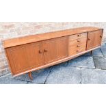 A 1960S TEAK SIDEBOARD fitted with four drawers flanked to one side with drop-down cupboard
