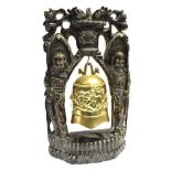 AN ORIENTAL BRASS BELL contained in a highly carved figural stand, inset with white metal, the