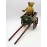 A threadbare vintage teddy, riding in a miniature polychrome-painted pine cart, measuring approx.